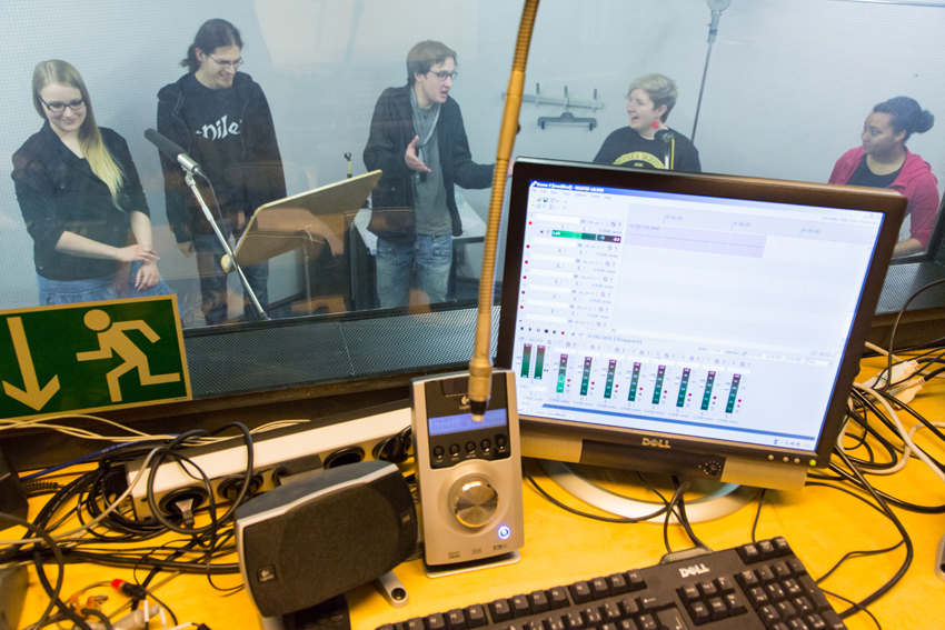 Ein Tonstudio von innen: Im Vordergrund steht ein Computer, eine Tastatur, Lautsprecher und ein Aufnahmegerät. In dem anderen Raum, im Hintergrund befinden sich fünf Personen an Mikrofonen und nehmen etwas auf.